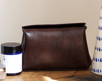 May include: A brown leather pouch, a jar of cream, and a white ceramic vase with a blue raindrop pattern, filled with dried pampas grass, are arranged on a wooden surface.