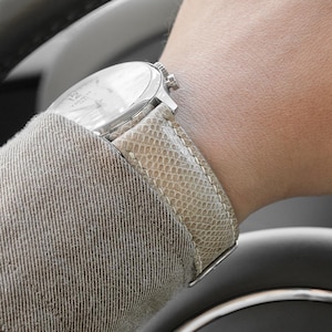 Peut inclure: Une montre-bracelet argentée avec un bracelet en peau de serpent de couleur claire est visible au poignet d'une personne. La main de la personne est sur un volant noir avec un emblème Jaguar argenté. La personne porte une veste grise.