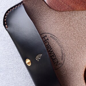 May include: A black leather glasses case with a gold button and the Horween logo. The case is open and the inside is brown leather.