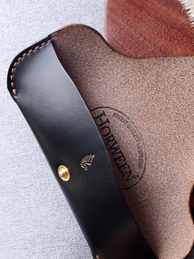 May include: A black leather glasses case with a gold button and the Horween logo. The case is open and the inside is brown leather.