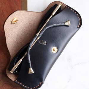 May include: A black leather sunglasses case with a brown stitch detail and a gold metal button. The case is open and a pair of sunglasses with a gold metal frame is inside.