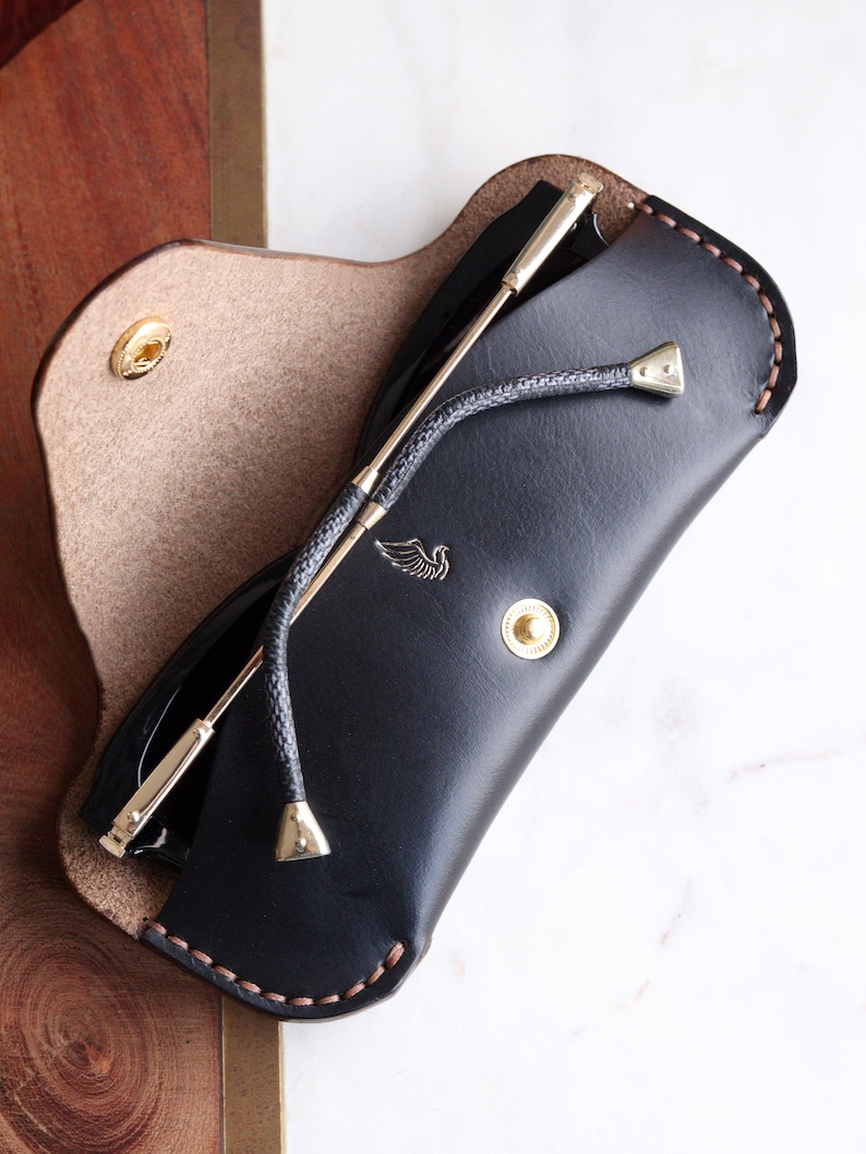 May include: A black leather sunglasses case with a brown stitch detail and a gold metal button. The case is open and a pair of sunglasses with a gold metal frame is inside.