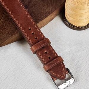 May include: A brown leather watch strap with stitching and a silver buckle. The strap is on a white surface.