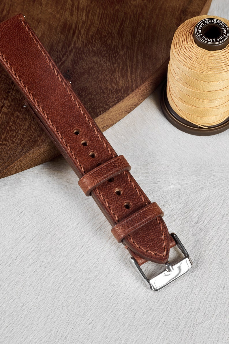 May include: A brown leather watch strap with stitching and a silver buckle. The strap is on a white surface.