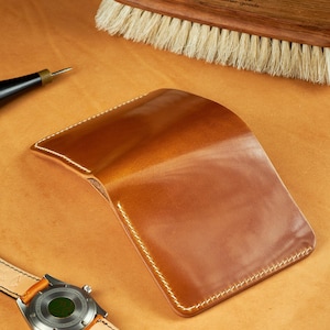 Może przedstawiać: Ręcznie wykonany, jasnobrązowy skórzany portfel z białymi przeszyciami, zegarek ze skórzanym paskiem i drewniana szczotka z napisem "Balzabany refined leather goods". Przedmioty znajdują się na jasnobrązowej powierzchni.