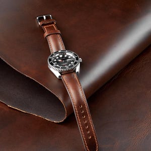 May include: A brown leather watch with a silver-colored bezel and a black watch face. The watch has a brown leather strap with a silver buckle. The watch is set against a brown leather background.