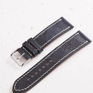 Puede incluir: Correa de reloj de cuero negro con costuras blancas. La correa tiene una superficie texturizada que se asemeja a la piel de avestruz.