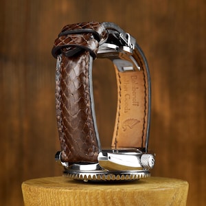 Op de afbeelding: Een close-up van een polshorloge met een bruine leren band met slangenpatroon. Het horloge heeft een zilverkleurige kast en een getextureerde lunette. Het horloge wordt op een houten standaard getoond.