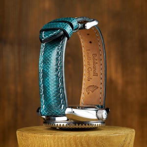 Puede incluir: Correa de reloj de cuero turquesa con costuras blancas, envuelta alrededor de una esfera plateada. La correa tiene un patrón de escamas. El reloj se muestra sobre un soporte de madera. La marca "Balabanoff Leather Goods" es visible.