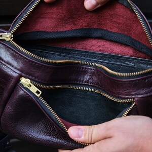 May include: A burgundy leather bum bag with a black zipper and a black lining. The bum bag is open and the lining is visible.