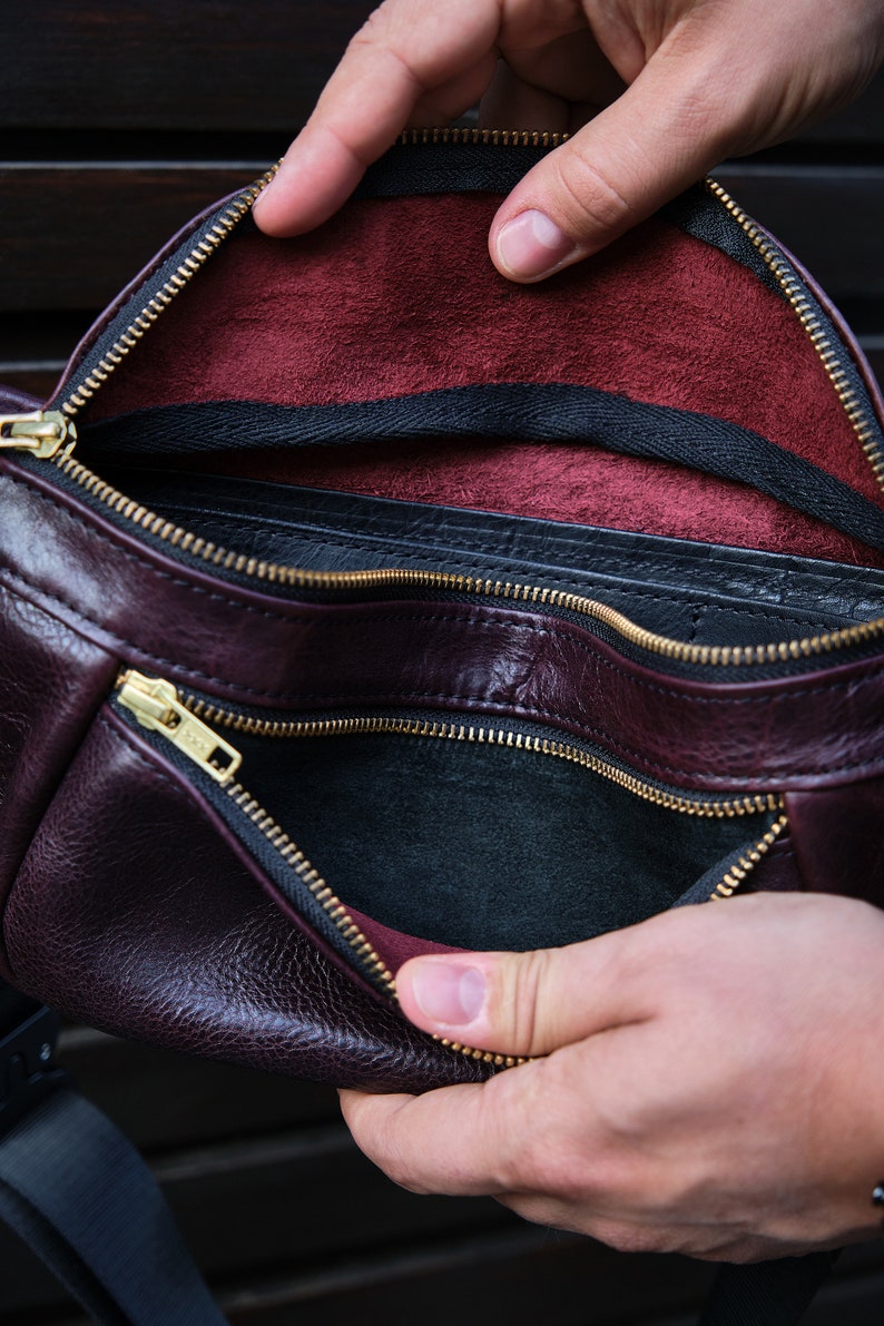 May include: A burgundy leather bum bag with a black zipper and a black lining. The bum bag is open and the lining is visible.