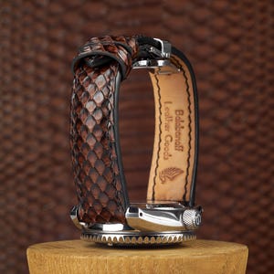 May include: A brown leather watch with a snakeskin pattern band. The watch has a silver-colored face and buckle. The watch is displayed on a wooden stand. The text "Balabanoff Leather Goods" is visible on the watch strap.