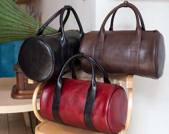 May include: Three leather duffel bags in black, brown, and red. The bags have a cylindrical shape and are arranged on a wooden chair.