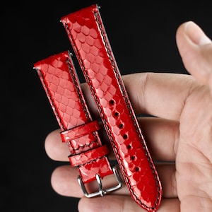 Puede incluir: Dos correas de reloj de cuero rojo con detalles de costura negra. Las correas están hechas de un material texturizado que se asemeja a la piel de serpiente.