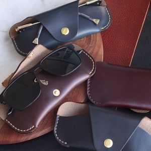 May include: Four leather eyeglass cases in different colors. Two are dark blue, one is brown, and one is burgundy. The cases are all closed and have a flap that folds over the glasses. The cases are all made of leather and have a stitched edge.