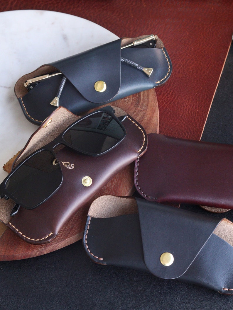 May include: Four leather eyeglass cases in different colors. Two are dark blue, one is brown, and one is burgundy. The cases are all closed and have a flap that folds over the glasses. The cases are all made of leather and have a stitched edge.