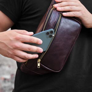 May include: A brown leather fanny pack with a zipper closure and a gold zipper pull. A smartphone is partially visible inside the pack.