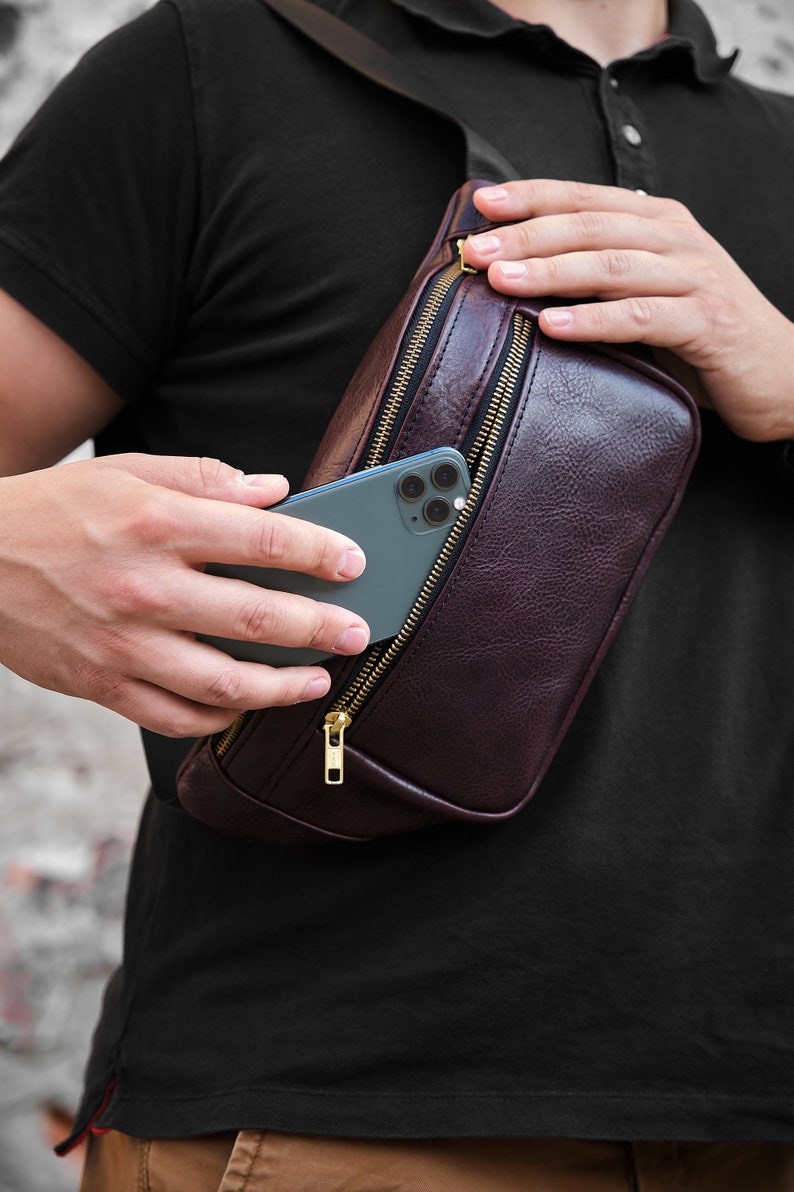 May include: A brown leather fanny pack with a zipper closure and a gold zipper pull. A smartphone is partially visible inside the pack.