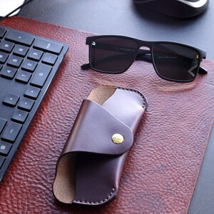 May include: A brown leather case for sunglasses with a gold button closure. The case is lying on a brown leather surface next to a pair of black sunglasses.