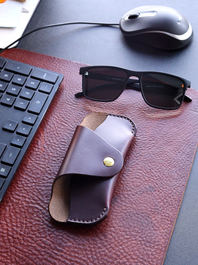 May include: A brown leather case for sunglasses with a gold button closure. The case is lying on a brown leather surface next to a pair of black sunglasses.