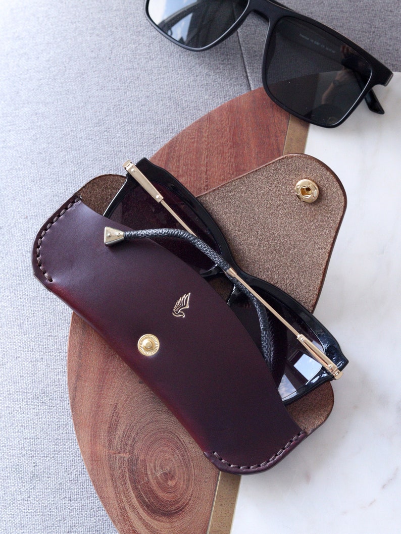 May include: A burgundy leather sunglasses case with a gold button and a small winged logo. The case is open and a pair of black sunglasses are inside.