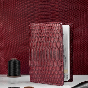 May include: A deep red passport holder featuring a textured snakeskin pattern with visible stitching along the edges. The holder is open, revealing white pages inside. A black spool of thread, three small metal coins, and a tool are nearby.