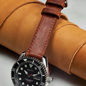 May include: A black and silver wristwatch with a brown leather band. The watch face has white markings and a date window. The watch is a limited edition automatic 300m.
