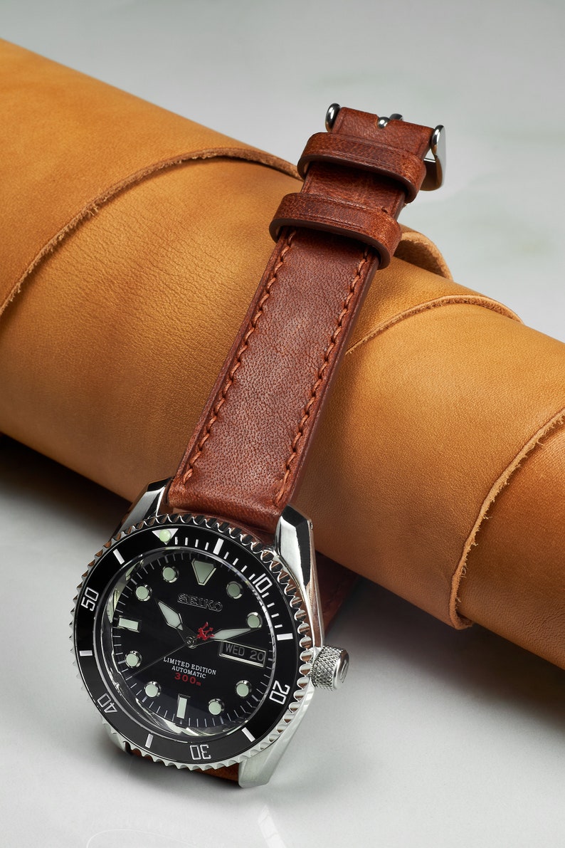 May include: A black and silver wristwatch with a brown leather band. The watch face has white markings and a date window. The watch is a limited edition automatic 300m.