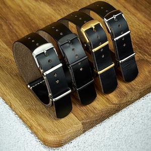 May include: Four black leather watch straps with different buckle finishes: silver, black, gold, and silver. The straps are arranged on a wooden surface, showcasing the variety of hardware options available for watch customization.
