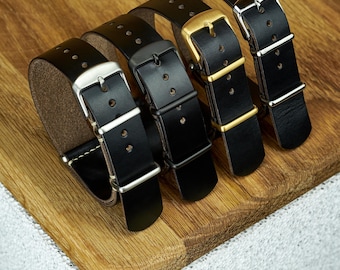May include: Four black leather watch straps with different buckle finishes: silver, black, gold, and silver. The straps are arranged on a wooden surface, showcasing the variety of hardware options available for watch customization.