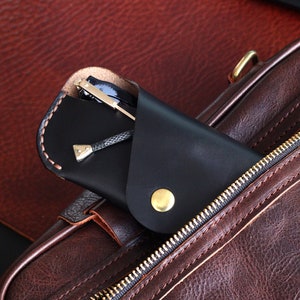 May include: A black leather glasses case with a gold button closure, tucked into a brown leather bag with a zipper.