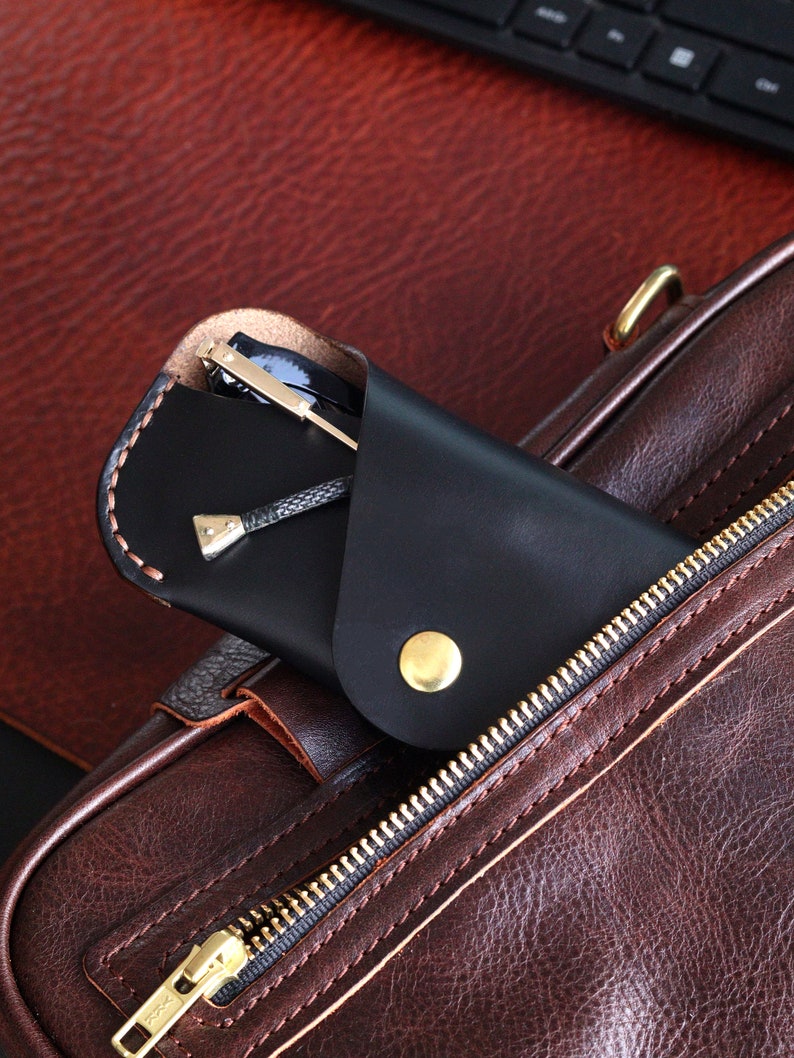 May include: A black leather glasses case with a gold button closure, tucked into a brown leather bag with a zipper.