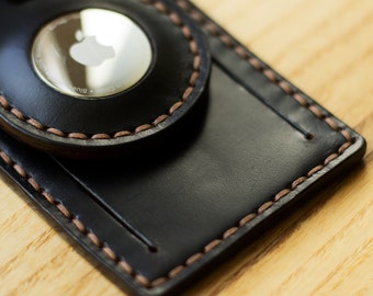 May include: A black leather luggage tag with a brown stitched border. The tag has a circular metal insert with the Apple logo and the text "Designed by Apple in California" in a silver font.