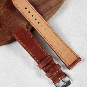 May include: Two brown leather watch straps with stitching. The strap on the left has a silver buckle. The strap on the right has the text "Baldane Fr Leather Goods" and a bird logo.