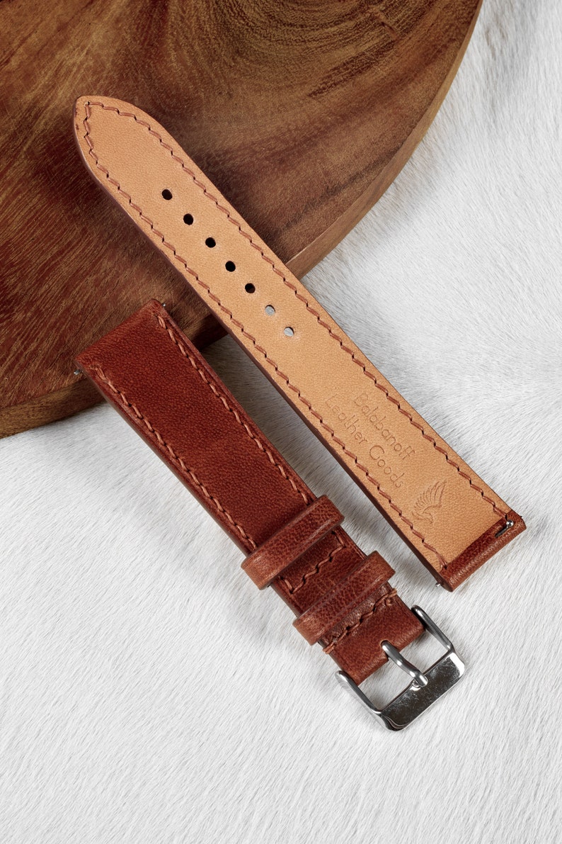 May include: Two brown leather watch straps with stitching. The strap on the left has a silver buckle. The strap on the right has the text "Baldane Fr Leather Goods" and a bird logo.