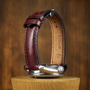 May include: A wristwatch with a burgundy leather band and a silver-toned watch face. The band is curved, showcasing the watch's design. The watch is displayed on a wooden stand, with the brand name "Balabanoff Leather Goods" visible.