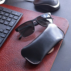 May include: Black leather glasses case with a stitched edge, next to a pair of black sunglasses and a black computer mouse on a brown leather desk mat.