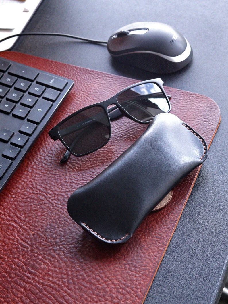 May include: Black leather glasses case with a stitched edge, next to a pair of black sunglasses and a black computer mouse on a brown leather desk mat.