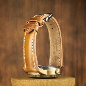 Op de afbeelding: Een close-up van een polshorloge met een bruinleren band en een goudkleurige wijzerplaat. De band is gestikt met witte draad en heeft de tekst "Balabanoff Leather Goods" aan de binnenkant. Het horloge wordt getoond op een houten standaard.