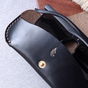 May include: Black leather sunglasses case with a gold button and a small winged logo. The case is open and a pair of black sunglasses are inside.