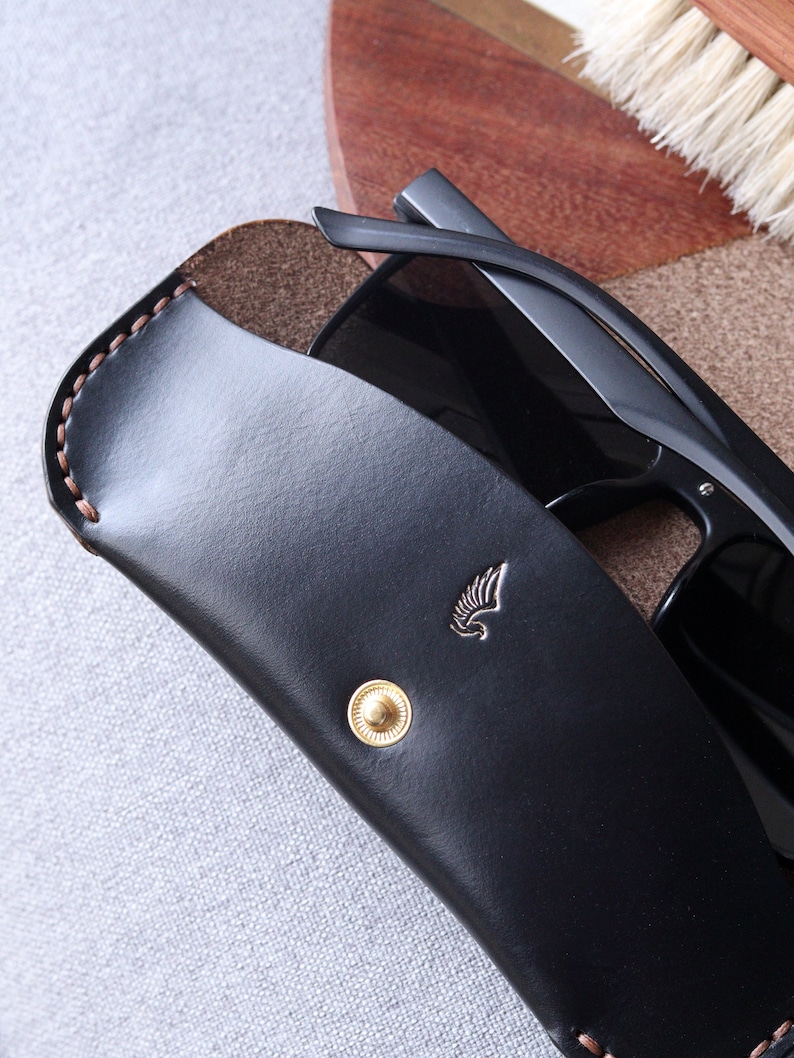 May include: Black leather sunglasses case with a gold button and a small winged logo. The case is open and a pair of black sunglasses are inside.