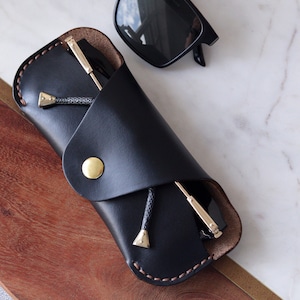 May include: A black leather glasses case with a brown stitching detail and a gold button closure. The case is folded over and a pair of black sunglasses are inside.