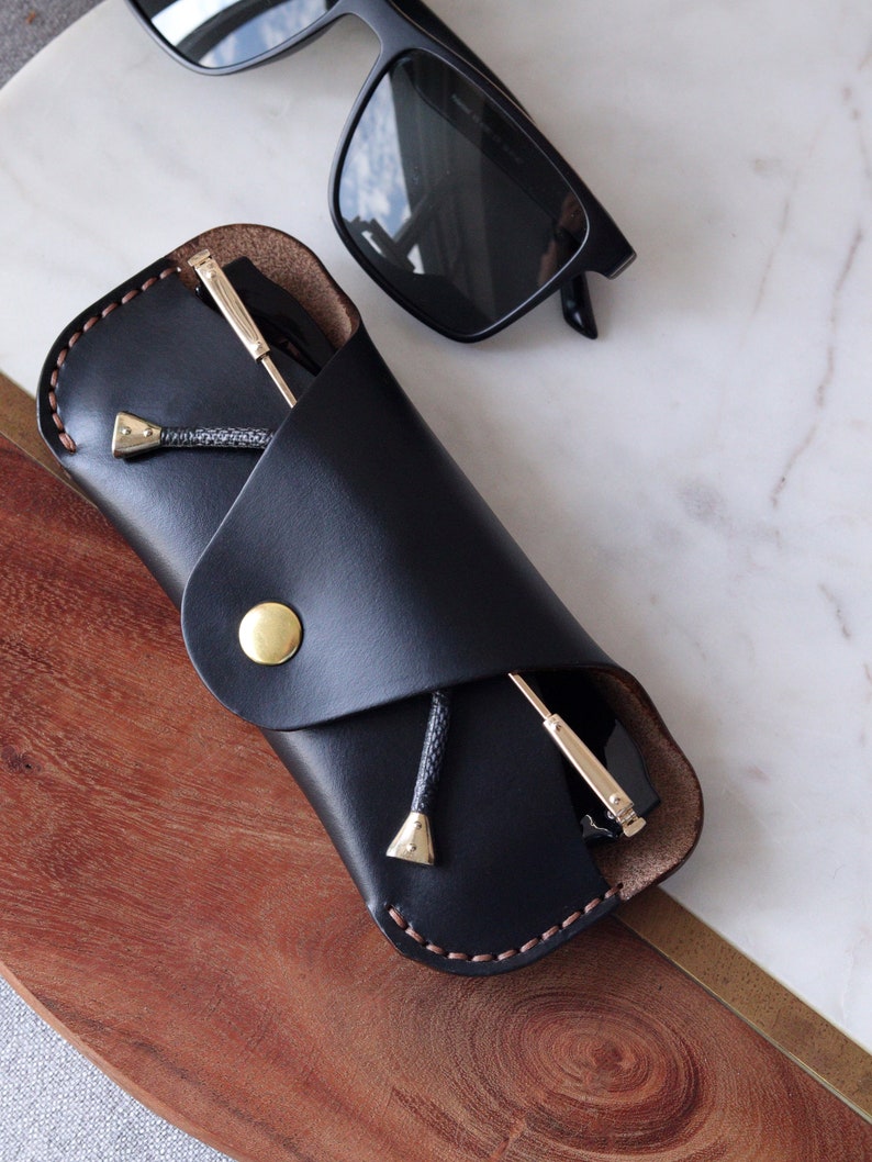 May include: A black leather glasses case with a brown stitching detail and a gold button closure. The case is folded over and a pair of black sunglasses are inside.