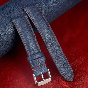 May include: Navy blue leather watch straps with red stitching. The straps have a textured surface and a silver-coloured buckle. The watch straps are displayed on a red surface.