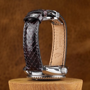 May include: A wristwatch with a dark burgundy, textured snakeskin-patterned leather strap. The inner side of the strap features the embossed text Balabanoff Leather Goods and a small logo. The watch case is made of polished silver-tone metal.