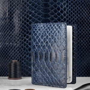 May include: A vertical passport holder featuring a deep blue, textured snakeskin pattern. The exterior displays a glossy finish with visible scale details. The interior contains white pages, and the item is positioned upright on a flat surface.