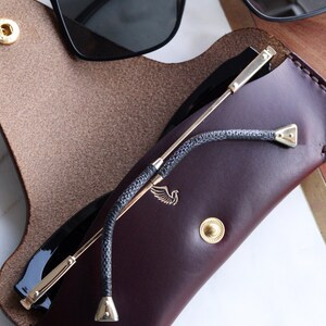 May include: A burgundy leather glasses case with a gold clasp and a gold metal strap. The case has a small bird logo on the front. A pair of black sunglasses is inside the case.