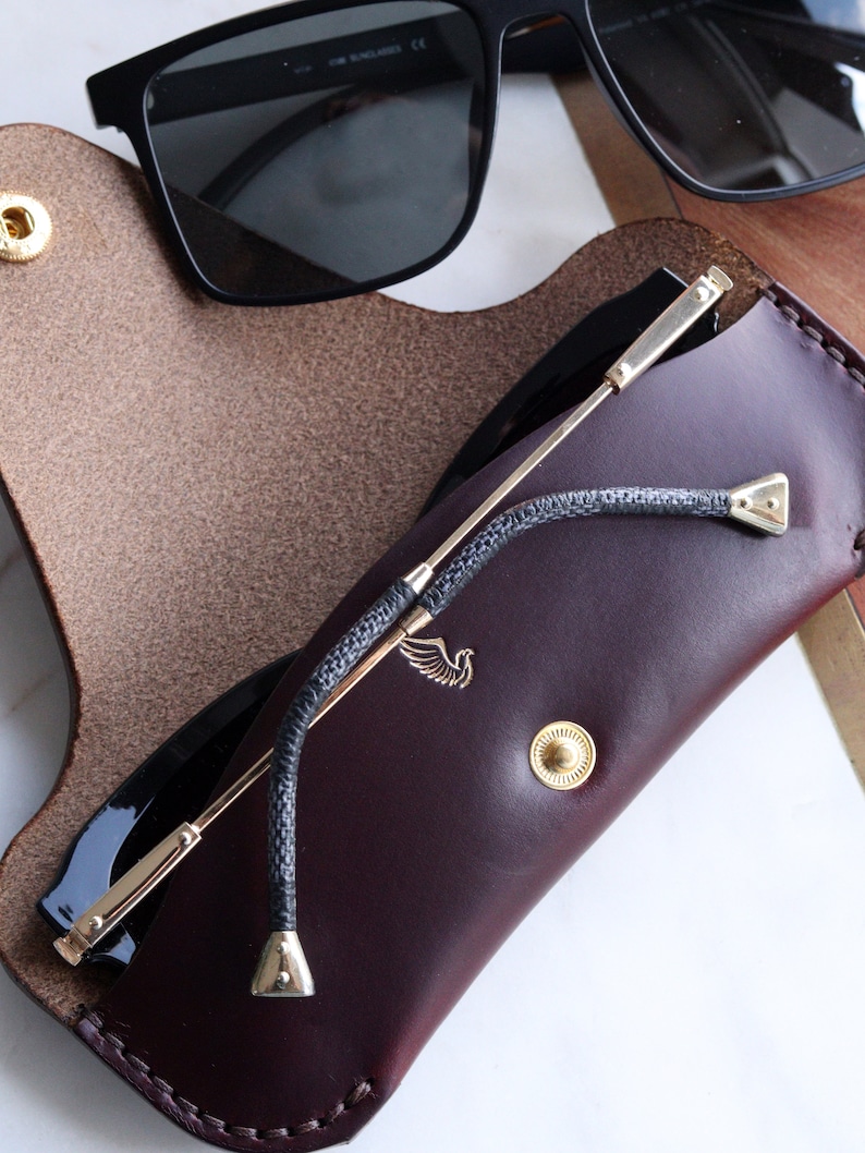 May include: A burgundy leather glasses case with a gold clasp and a gold metal strap. The case has a small bird logo on the front. A pair of black sunglasses is inside the case.
