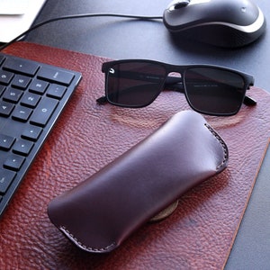 May include: A brown leather case for sunglasses, with a pair of black sunglasses on a brown leather desk surface.
