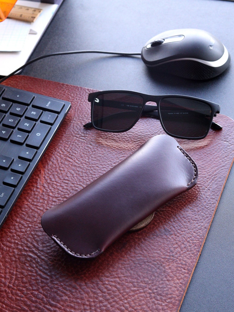 May include: A brown leather case for sunglasses, with a pair of black sunglasses on a brown leather desk surface.
